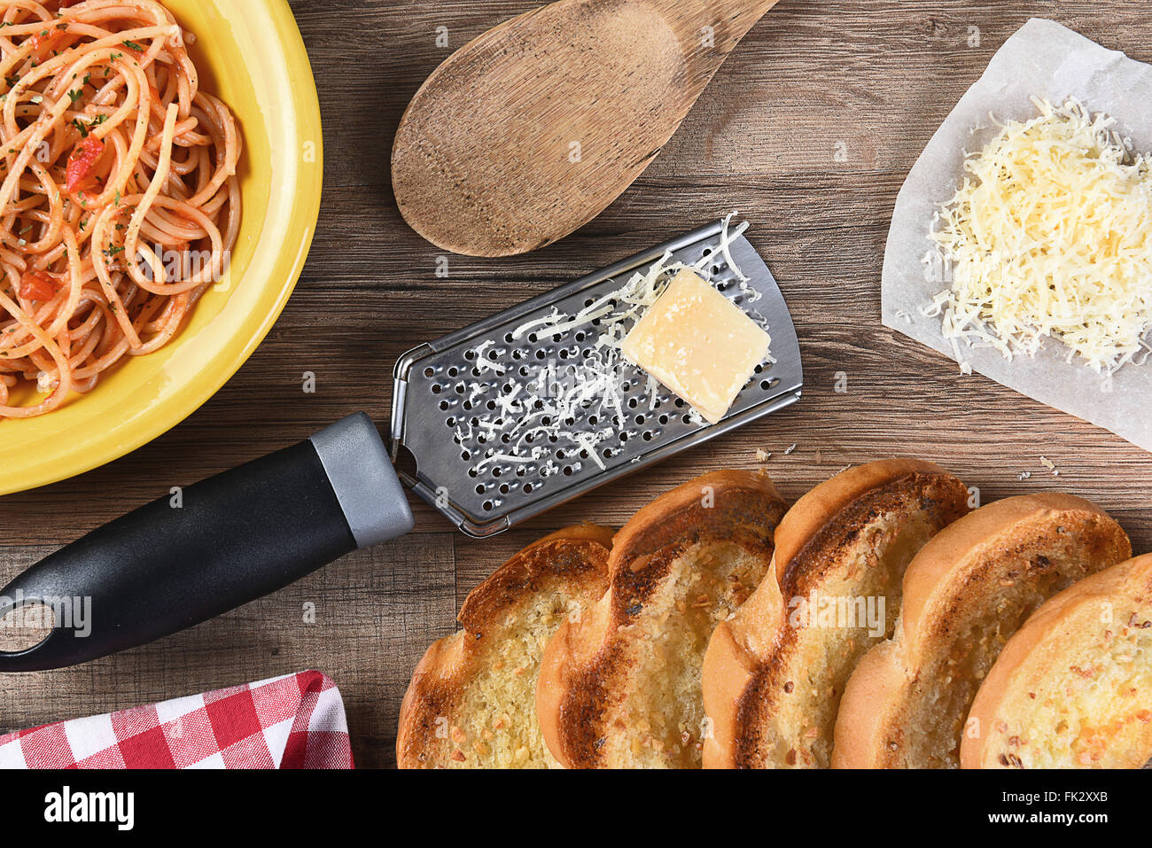 Draufsicht auf einen Küchentisch mit einem Teller Spaghetti, Knoblauchbrot, geriebenen Parmesan, Reibeisen, hölzerne Löffel, eine rote ch Stockfoto