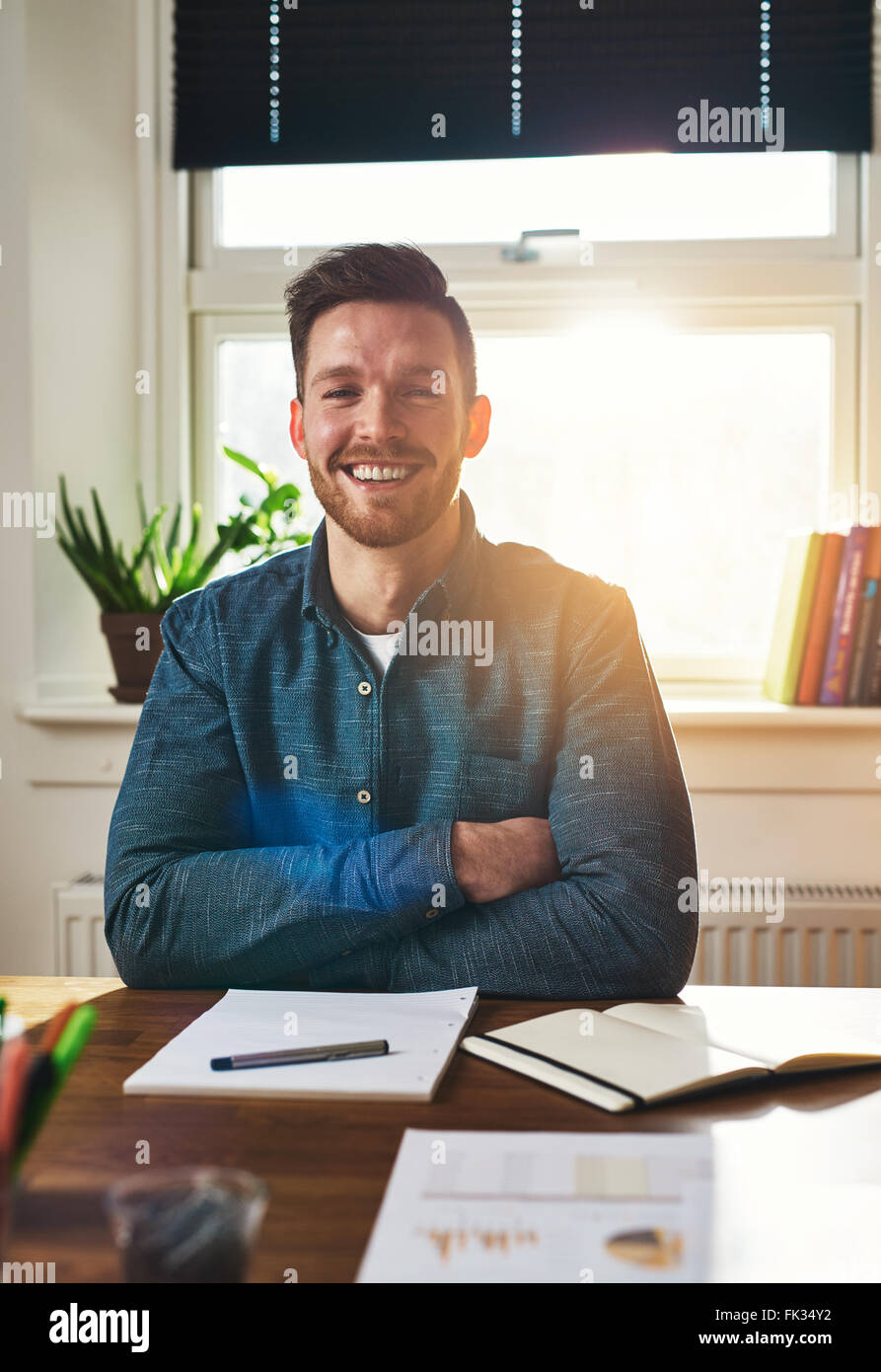 Lächelnd, glücklichen erfolgreichen Geschäftsmann sitzt an seinem Schreibtisch mit verschränkten Armen lachend in die Kamera mit Papierkram auf dem Schreibtisch im f Stockfoto