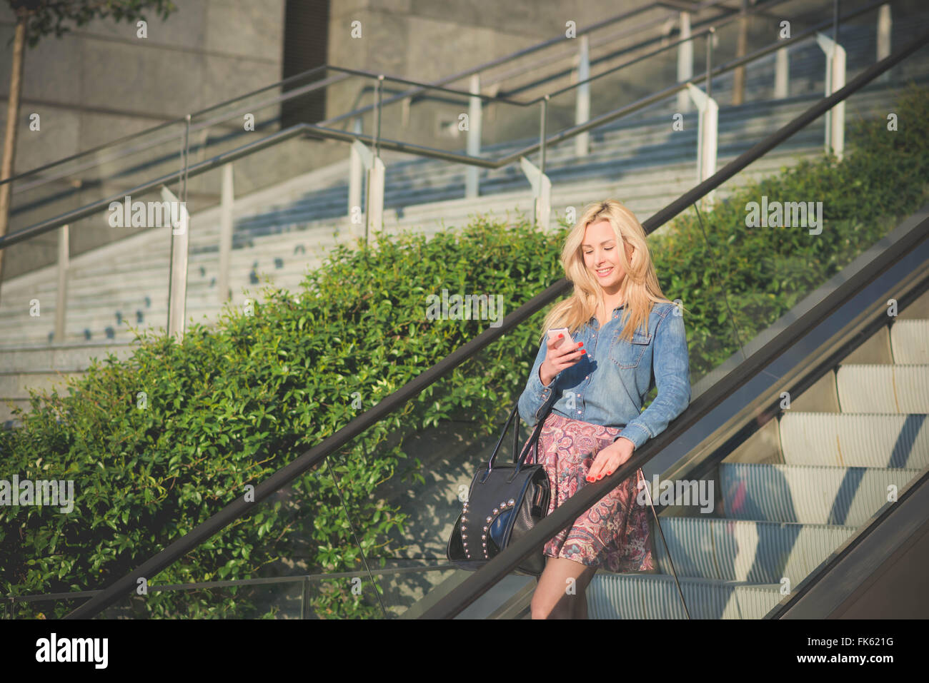 Knie-Figur des jungen schönen kaukasischen blonde Mädchen auf der Rolltreppe mit einem Smartphone suchen th Bildschirm Lächeln - Technologie, Kommunikation, soziale Netzwerk-Konzept Stockfoto