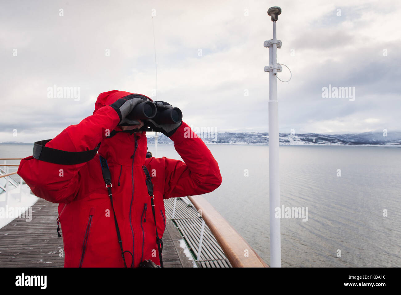 Ein Mann in einem roten Mantel verwendet Fernglas auf dem Deck eines Kreuzfahrtschiffes in Arktis Norwegen. Stockfoto