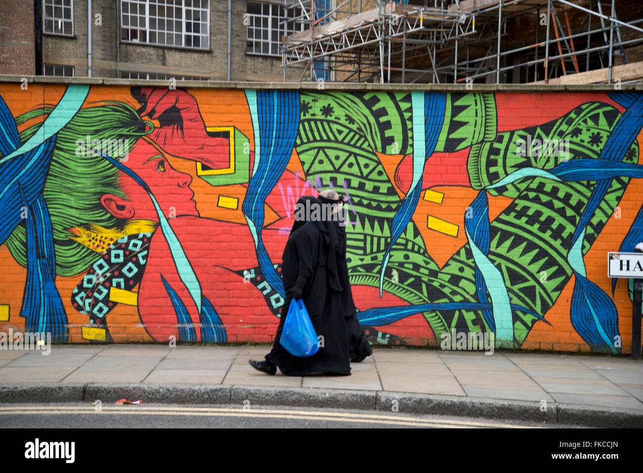 Hanbury Street, an der Brick Lane. Tower Hamlets. Zwei Frauen tragen schwarze Mäntel und Schleier gehen vorbei an einem bunten Wandgemälde. Stockfoto