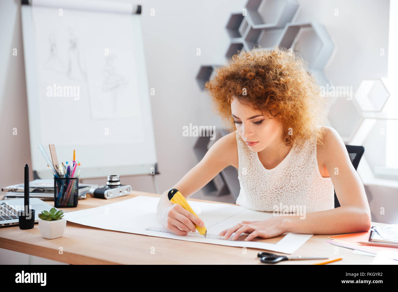 Schöne fokussierte junge Frau Fotograf Cuttung Blatt Papier mit Briefpapier Messer und Lineal auf ihrem Arbeitsplatz Stockfoto