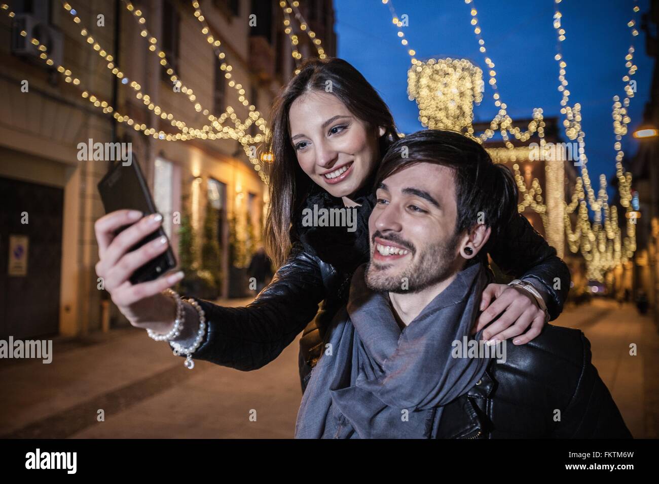 Junger Mann geben junge Frau Huckepack mit Smartphone zu Selfie, Lächeln Stockfoto