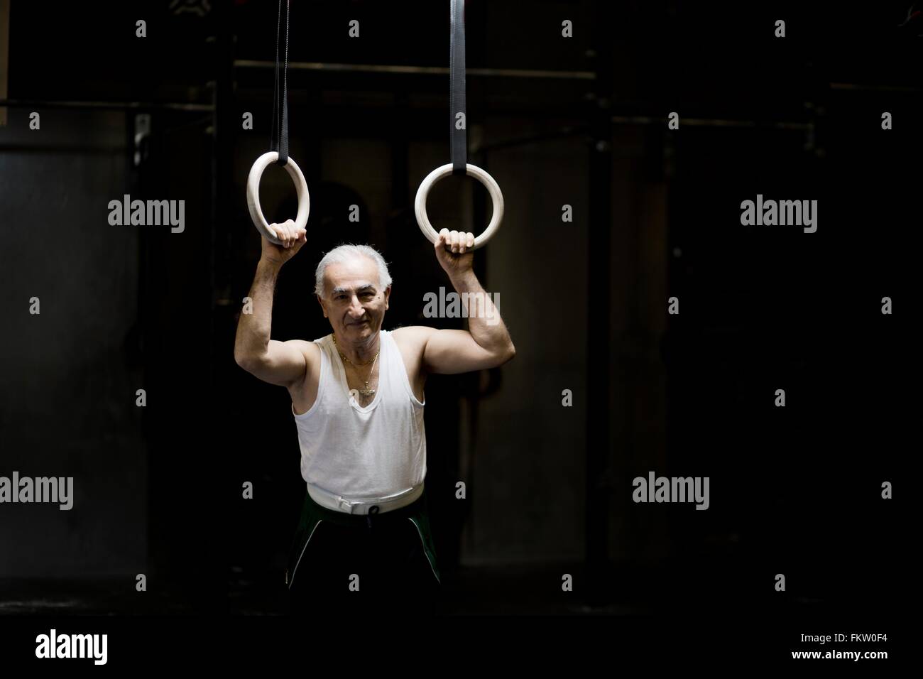 Porträt von grauen Haaren senior Mann, hält Gym Ringe in dunklen Turnhalle Stockfoto
