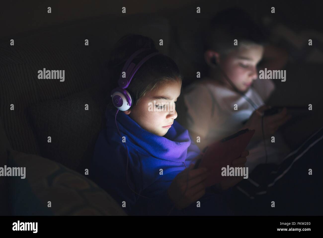 Jungen und Mädchen nebeneinander auf dem Sofa tragen Kopfhörer mit digitalen Tabletten Stockfoto