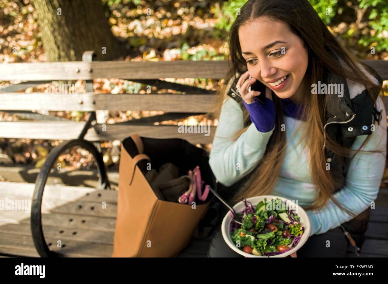 Junge Frau sitzt am Park Bench im Chat auf Smartphone beim Mittagessen Salat essen Stockfoto