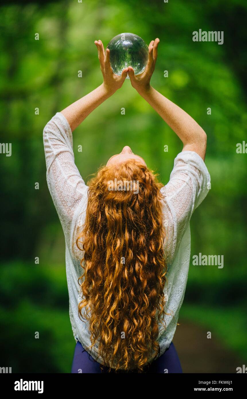 Rückansicht des junge Frau mit langen roten Haaren hält Kristallkugel im Wald Stockfoto