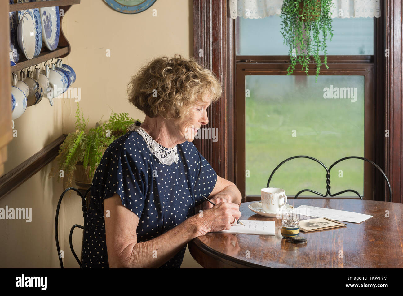 In einer historischen Szene aus dem 30er oder 40er Jahren schreibt eine attraktive ältere Frau einen Brief, als sie an einem Tisch am Fenster sitzt. Stockfoto
