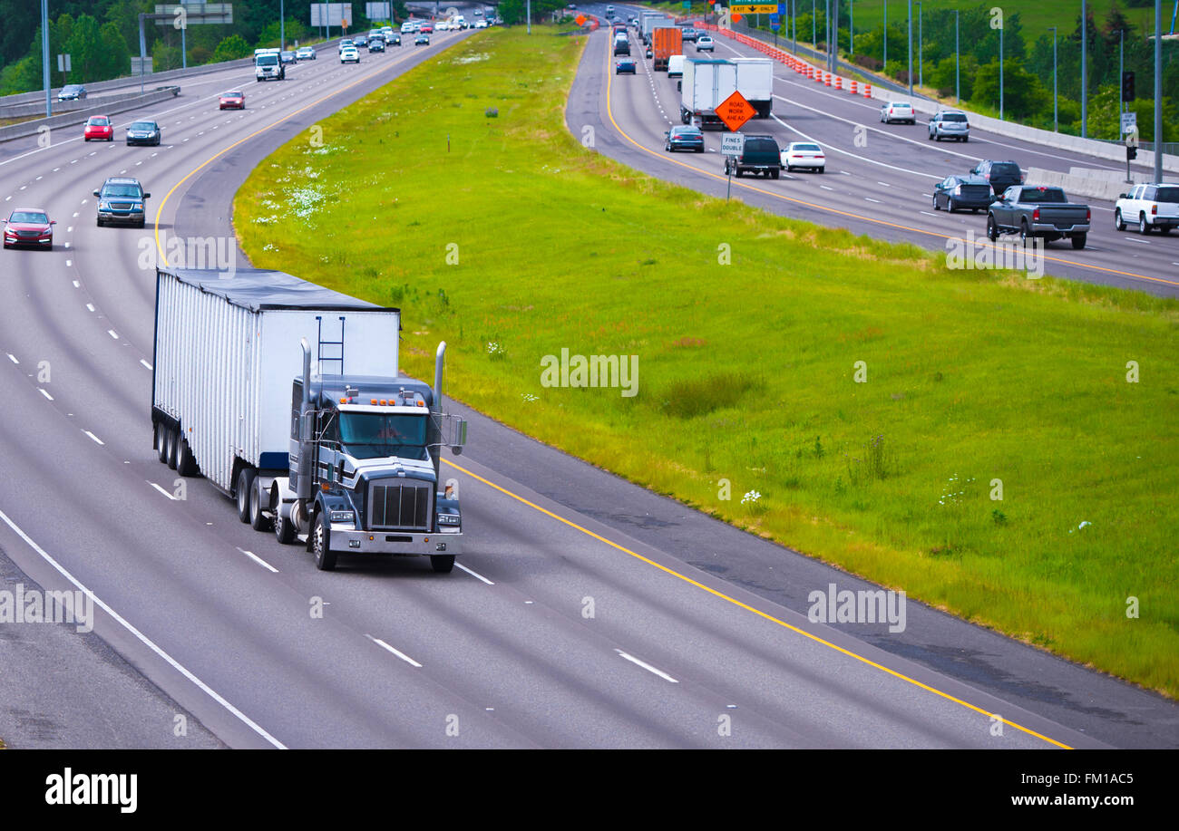 Eine breite Autobahn mit unbefahrbaren Gras gegenüberliegenden Seiten mit einer klassischen big Rig LKW Verkehr und Bulk Auflieger im Vordergrund. Stockfoto