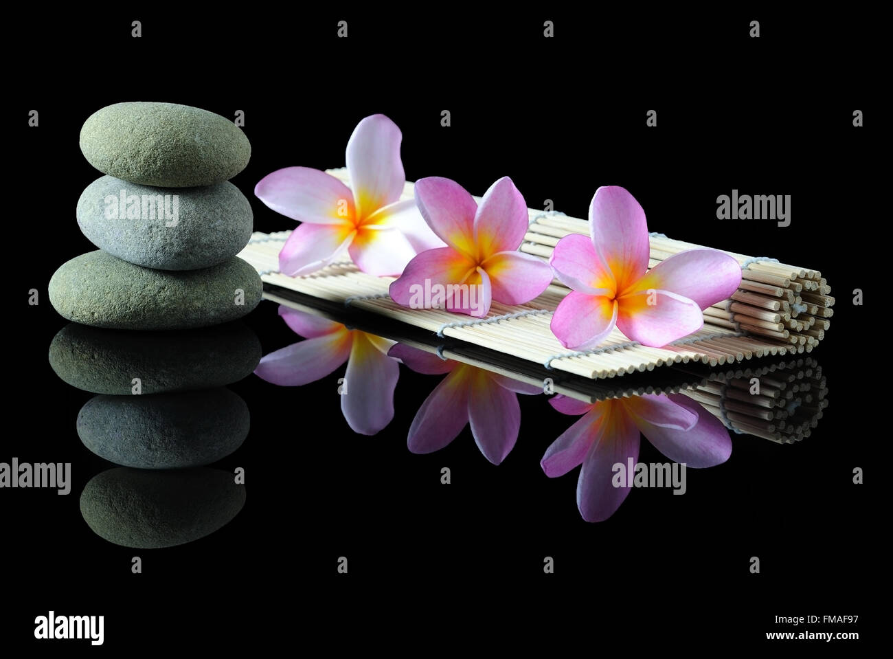 Wellness-, Beauty- und Wellness-Konzept - gestapelt Zen Steinen Frangipani-Blüten auf einer Bambusmatte mit Reflexion, dunklen Hintergrund. Stockfoto