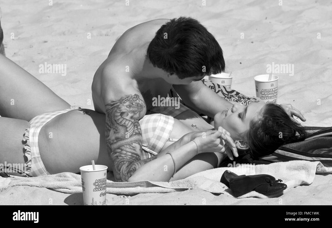 Reportageaufnahme attraktives Paar, Mann mit Tattoos, Frau im Bikini, Tassen trinken, liegen am Strand, Zuneigung, Liebe Stockfoto