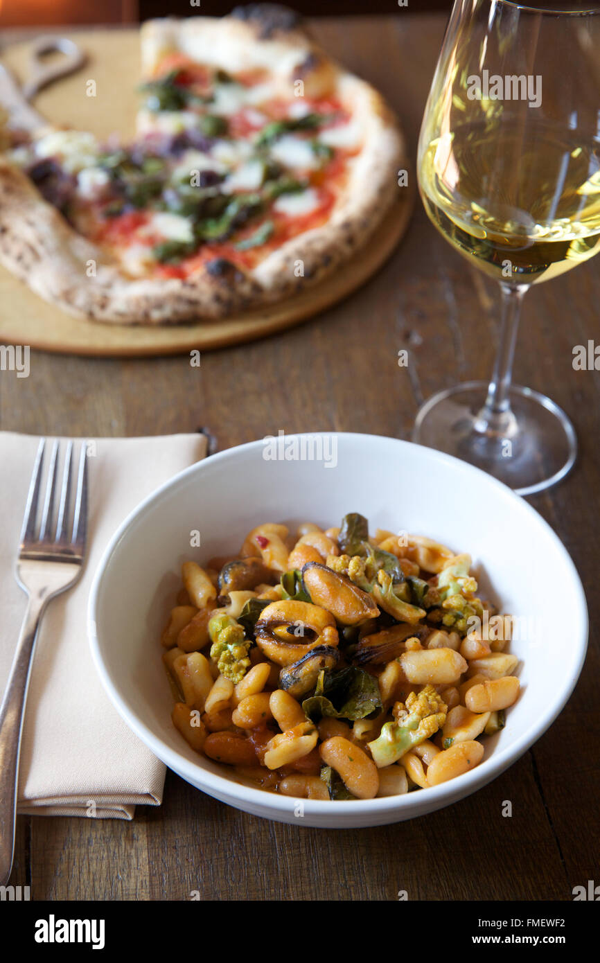 Eine Schale mit weißen Bohnen, Romanesco und Muscheln-Suppe mit Weißwein und eine Pizza auf dem Tisch hinter sich. Stockfoto