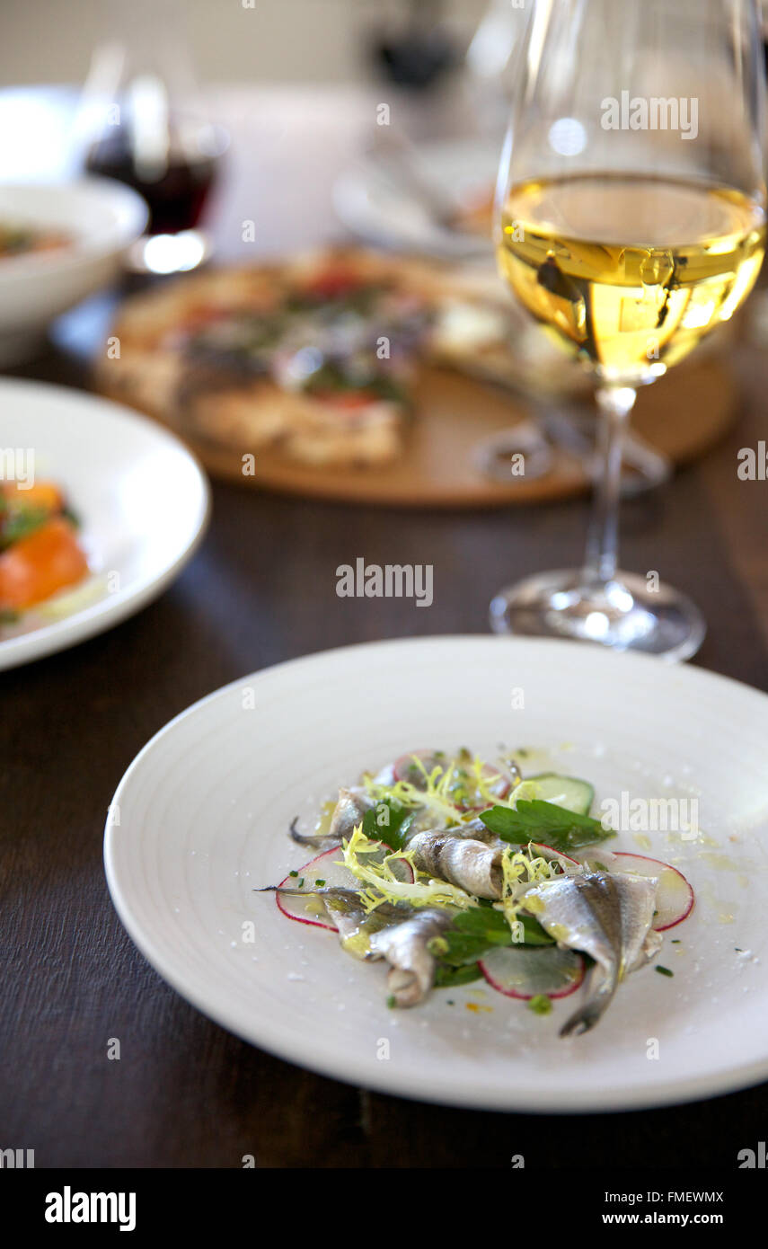 Ein Crudo Teller Sardinen mit Radieschen & Grüns, ein Glas Weißwein und andere Platten der umliegenden Essen zubereitet. Stockfoto
