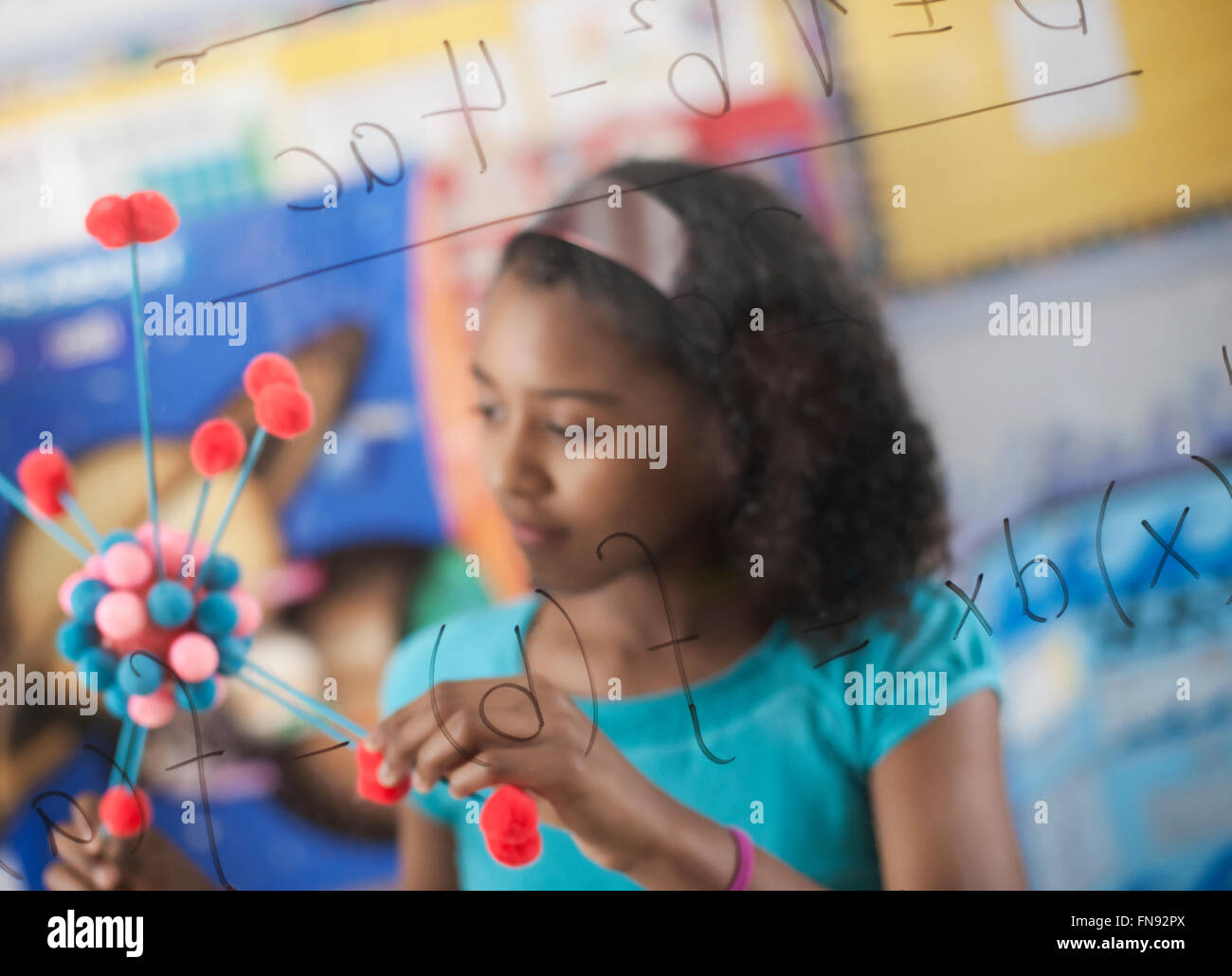 Ein Mädchen hält eine molekulare Struktur und mit Blick auf ein Brett von Gleichungen und Formeln im Klassenzimmer. Stockfoto