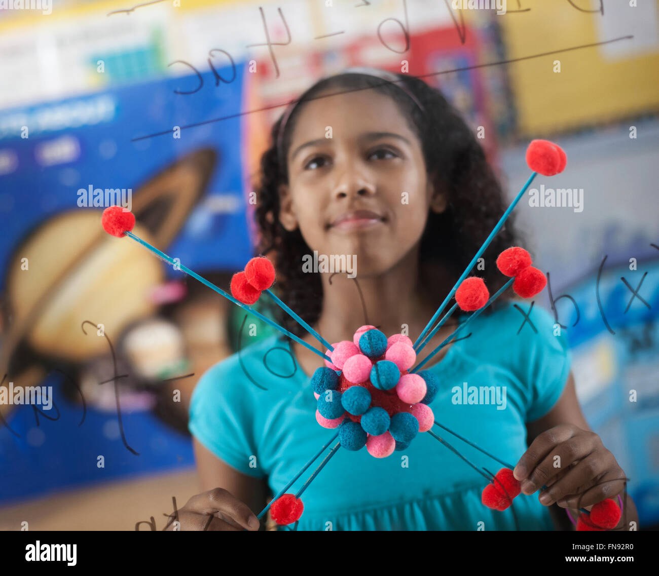 Ein Mädchen hält eine molekulare Struktur und mit Blick auf ein Brett von Gleichungen und Formeln im Klassenzimmer. Stockfoto