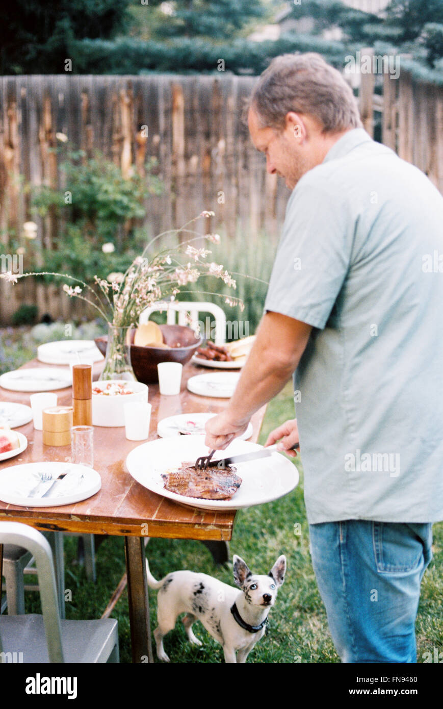 Ein Mann schnitzen Fleisch an einem Familienessen im Garten, beobachtet von einem kleinen Hund unter dem Tisch. Stockfoto