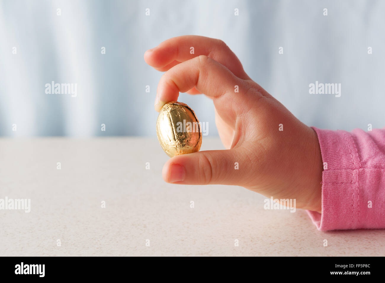 Des Kindes Hand hält ein Schokoladen Osterei verpackt in goldener Folie zwischen zwei Fingern mit Textfreiraum Stockfoto