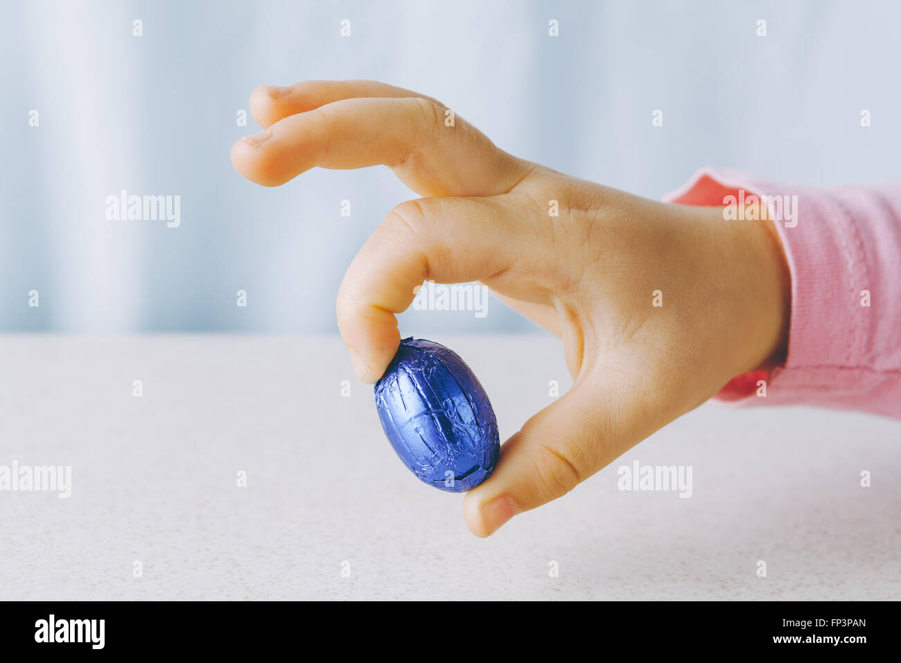Des Kindes Hand hält ein Schokoladenei Ostern eingehüllt in blaue Folie zwischen zwei Fingern mit Textfreiraum Stockfoto