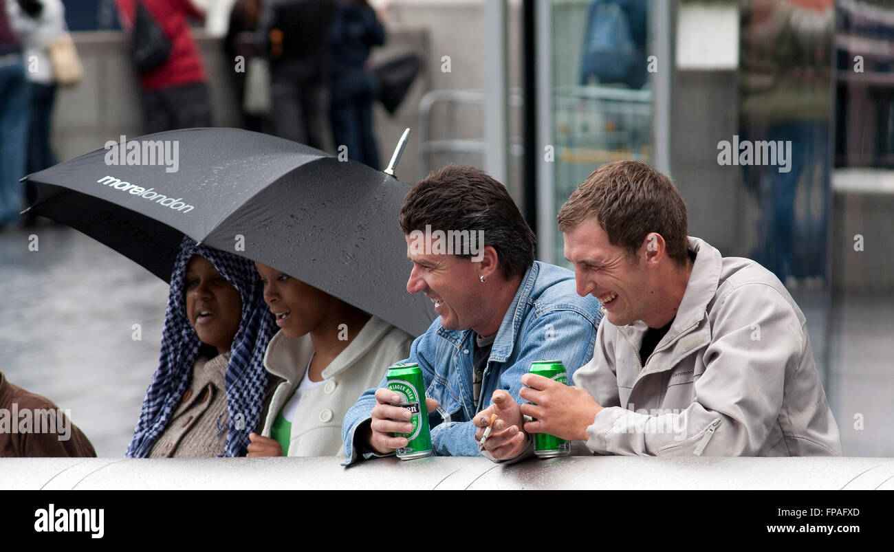 zwei Lachen männliche Touristen beobachten Straßenunterhaltung im Regen und Biertrinken London, UK Stockfoto
