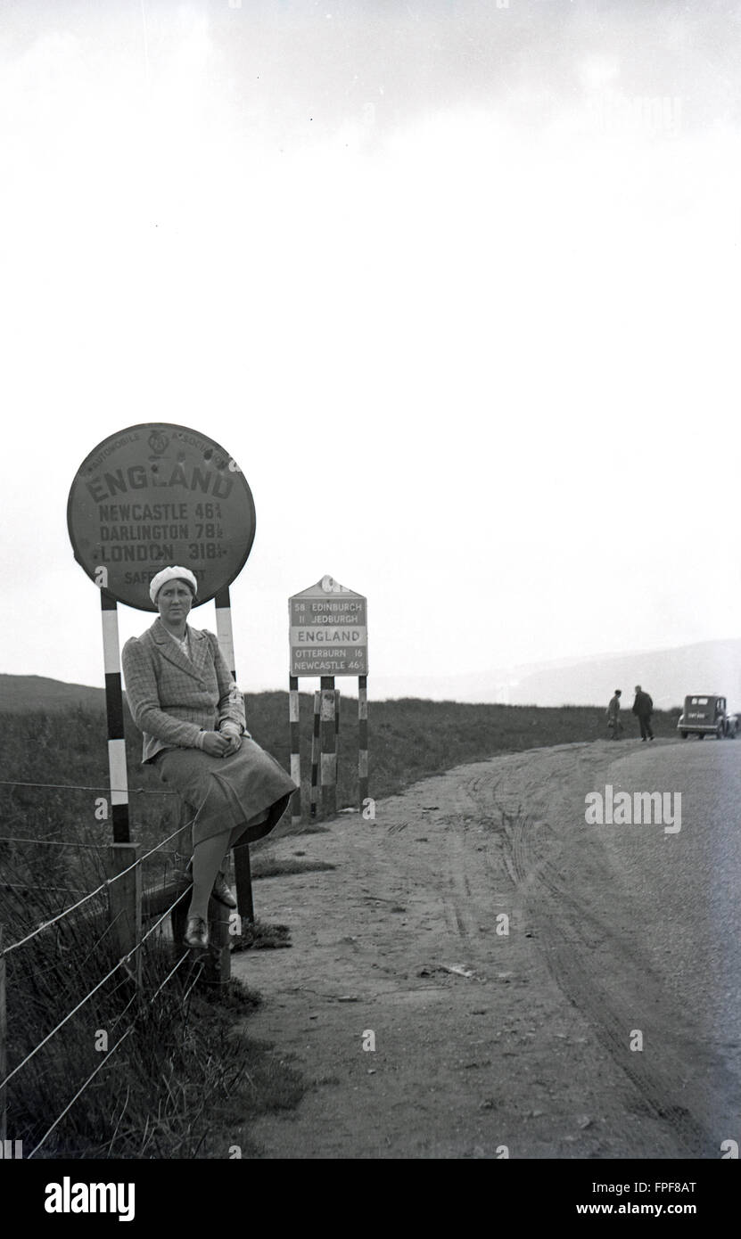 1930er Jahre historische, Lady sitzen am Straßenrand an einem Zaun vor der AA melden für England - Newcastle, Darlington und London (318 1/4 Meilen) - an der Grenze zu Schottland. Stockfoto