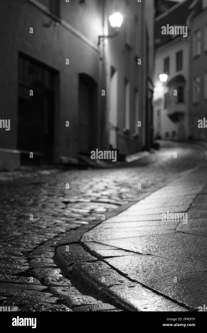 Beleuchtete Nacht Stadtstraße der alten europäischen Stadt. Unscharfe schwarz-weiß Bild mit Fokus auf Vordergrund Pflaster Stockfoto