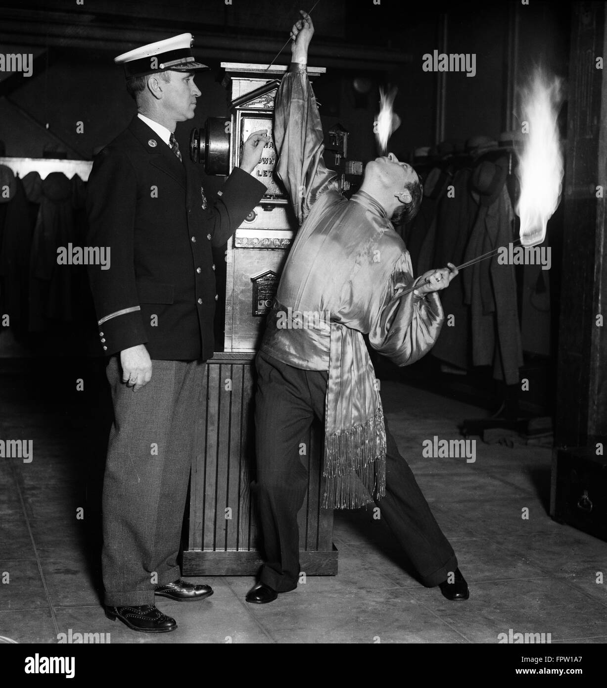 1930ER JAHRE MANN FEUER ESSER ZIRKUSKÜNSTLER FEUER ZU SPEIEN UND HALTEN FLAMMENDE WAND STEHEND FEUERALARM KASTEN MIT FEUERWEHRMANN ASSISTENT Stockfoto