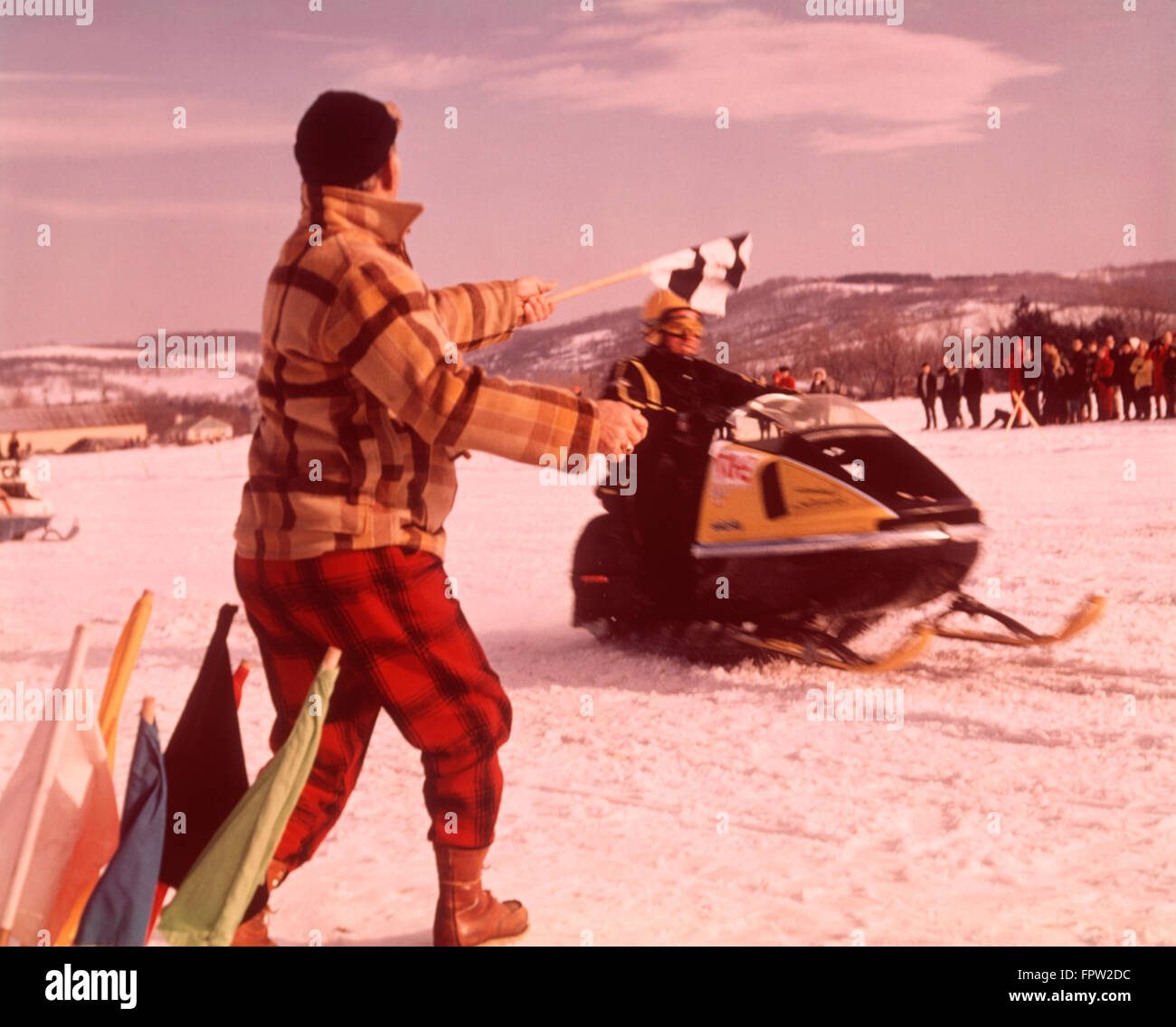 1960S 1970S SCHNEEMOBIL RENNEN MANN BEAMTER IN GELB ROT KARIERTEN WOLLE SCHNEE ANZUG WEHENDE ZIELFLAGGE KARIERTEN GEWINNER Stockfoto