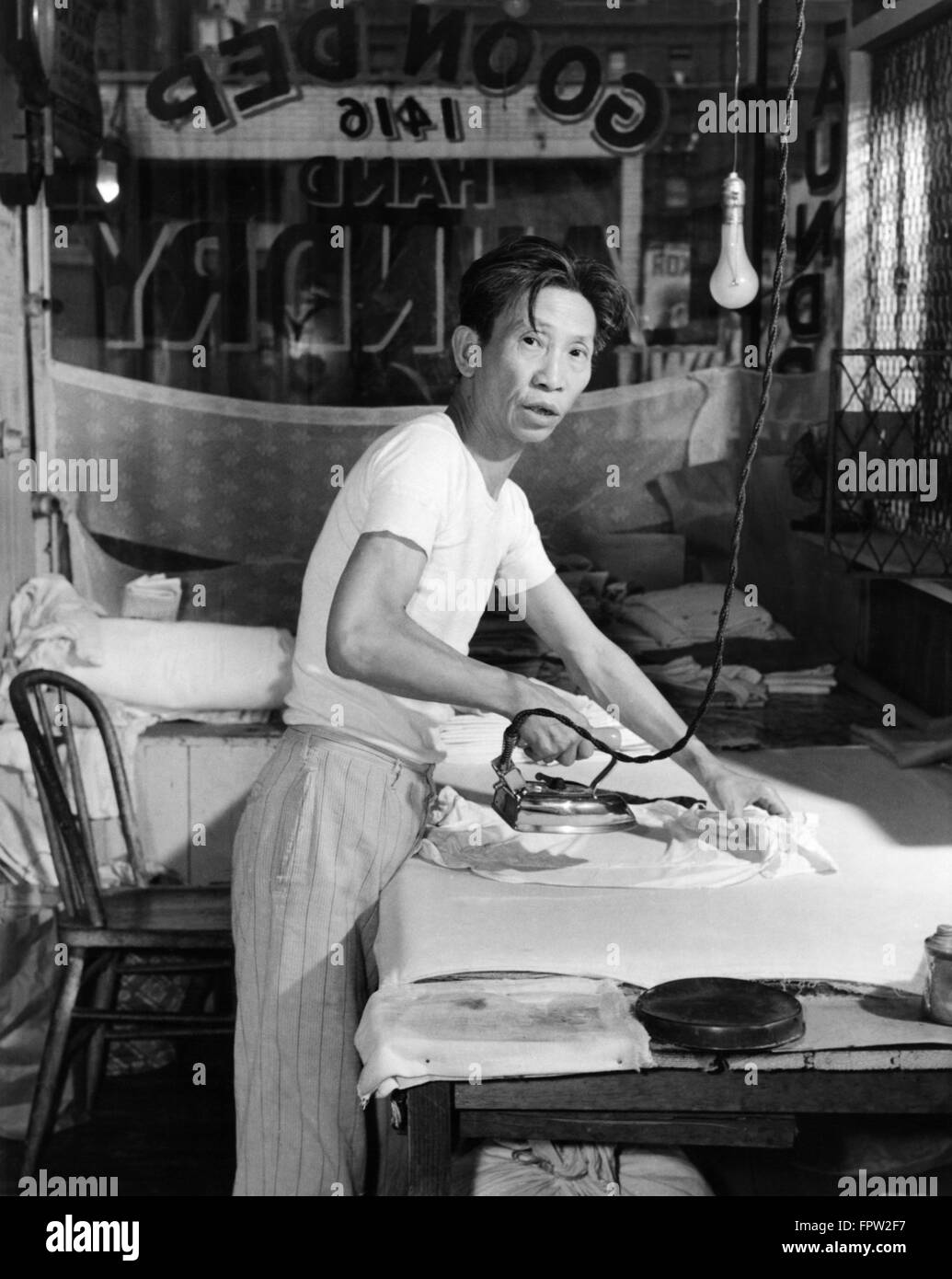 1920ER JAHRE CHINESE LAUNDRY MAN LOOKING AT KAMERA BÜGELN KLEIDUNG AN BORD Stockfoto