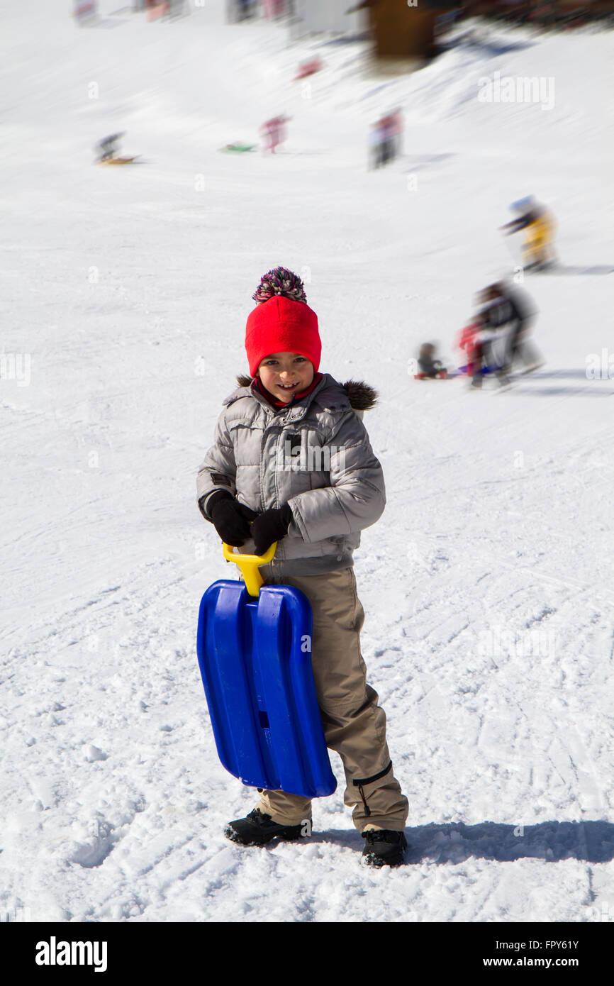 Kind im Schnee mit blauem Schlitten und Hut mit Pompons Stockfoto