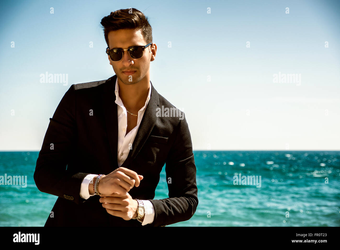 Schönen jungen Mann im klassischen Anzug am Strand halten Sonnenbrille beim wegschauen. Wellen des Meeres im Hintergrund Stockfoto