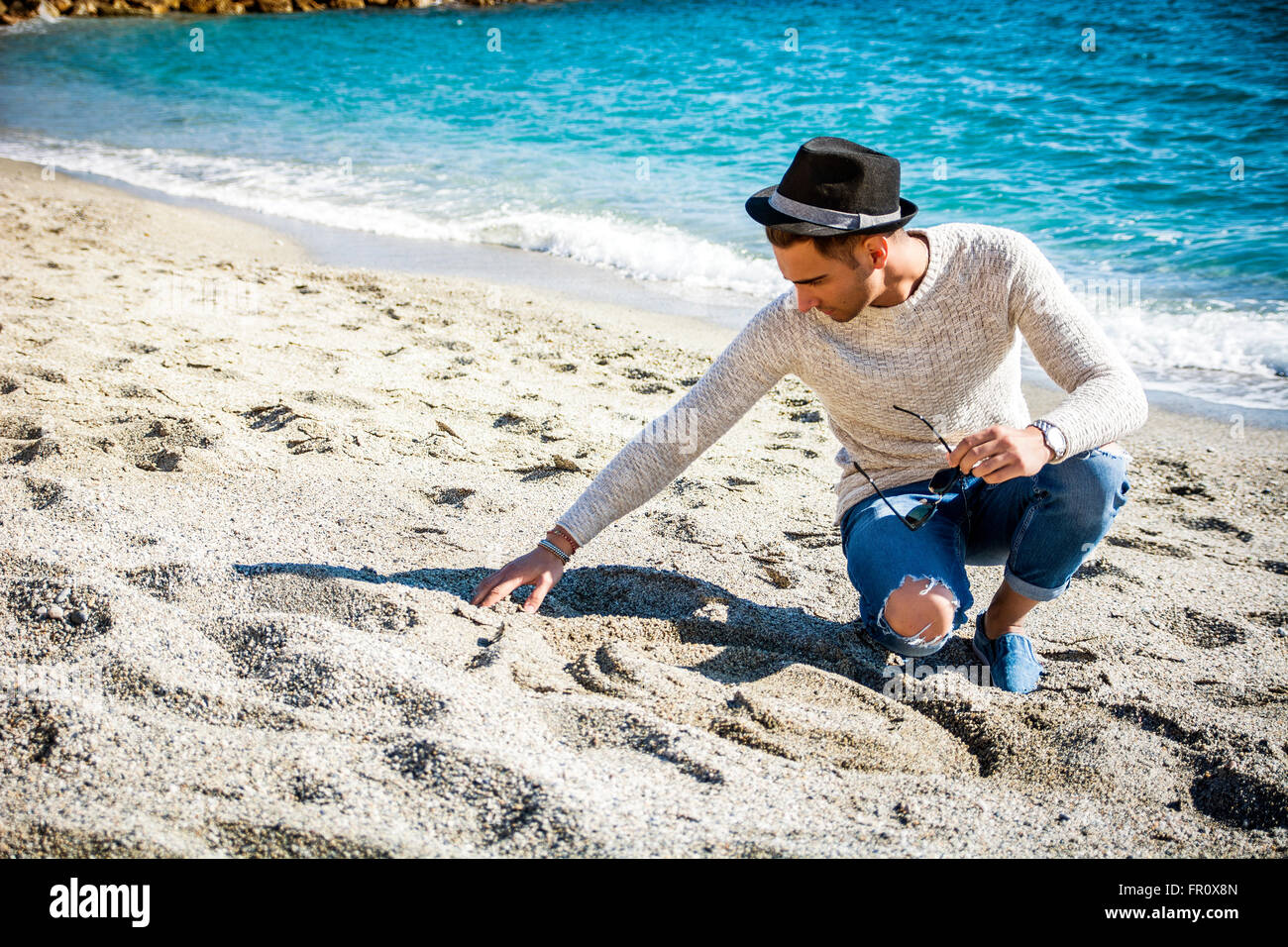 Junger Mann im schwarzen Hut sitzt am Strand und Sand berührt, während wegschauen Stockfoto