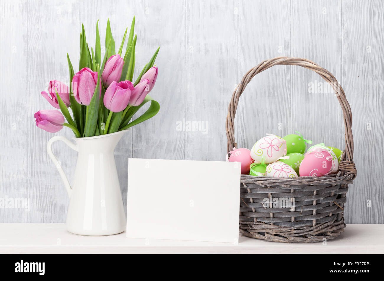 Ostereier in Baasket, Grußkarte und rosa Tulpen Strauß auf Regal vor Holzwand. Zeigen mit textfreiraum an Stockfoto