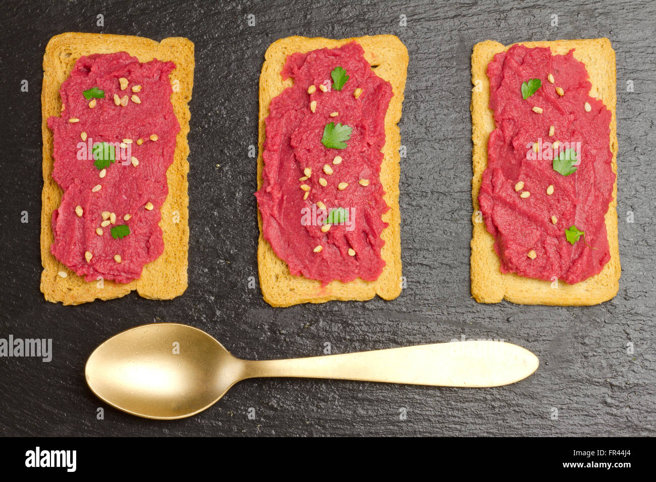 Drei Scheiben Brot mit Rüben Hummus und einem goldenen Löffel auf dem Schiefer Teller Stockfoto