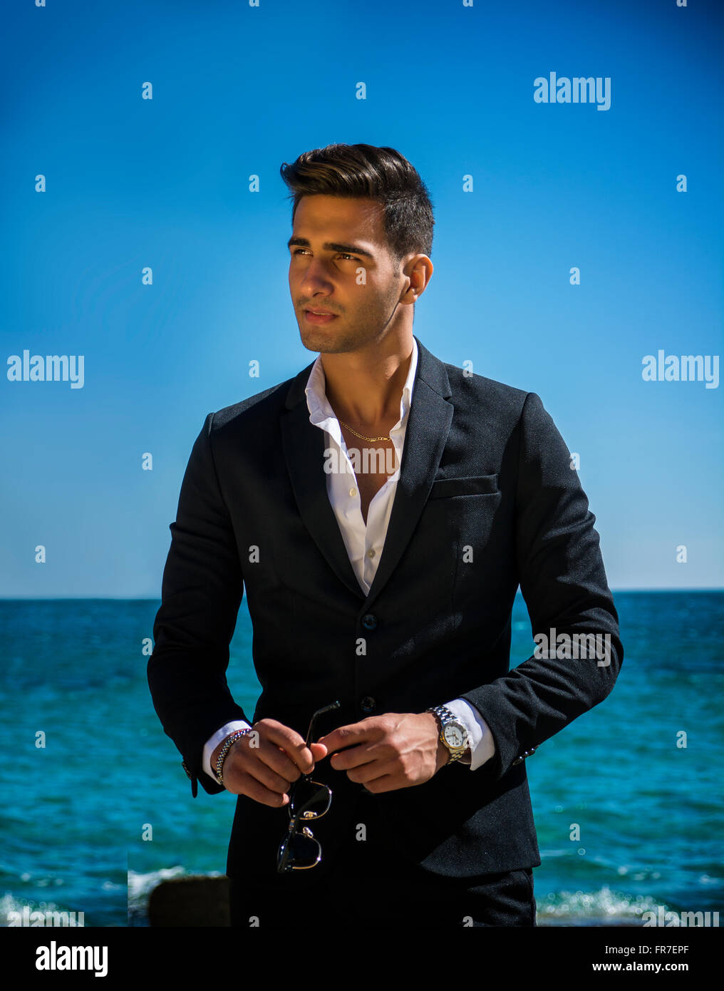 Schönen jungen Mann im klassischen Anzug am Strand halten Sonnenbrille beim wegschauen. Wellen des Meeres im Hintergrund Stockfoto