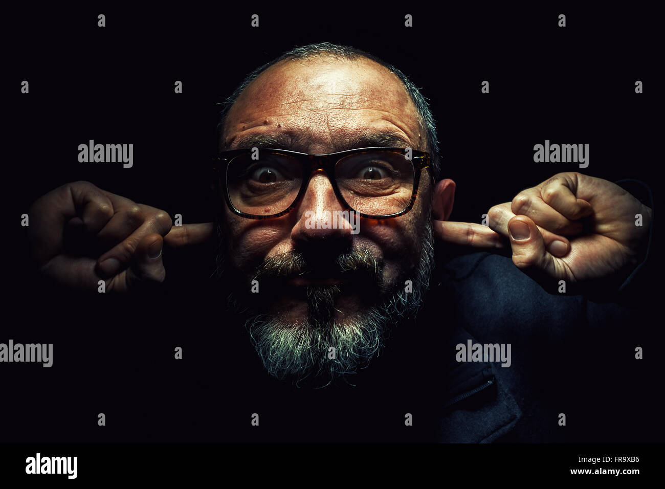 Porträt eines älteren Mannes mit Brille, mit den Fingern in den Ohren, Augen weit geöffnet. Stockfoto