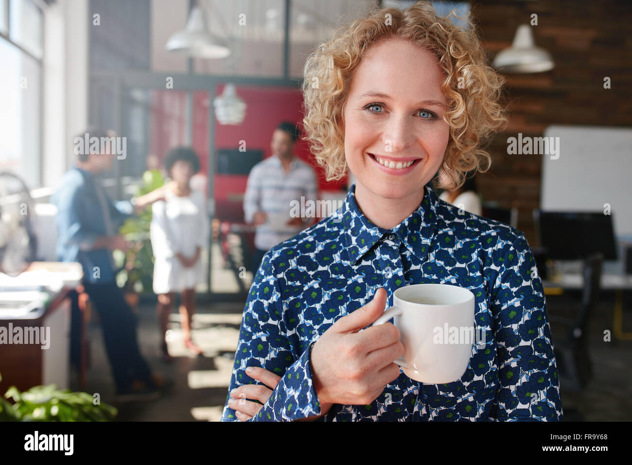 Porträt der glückliche junge Geschäftsfrau hält eine Tasse Kaffee, Blick in die Kamera und lächelnd. Sie steht in ihrem Büro mit Stockfoto