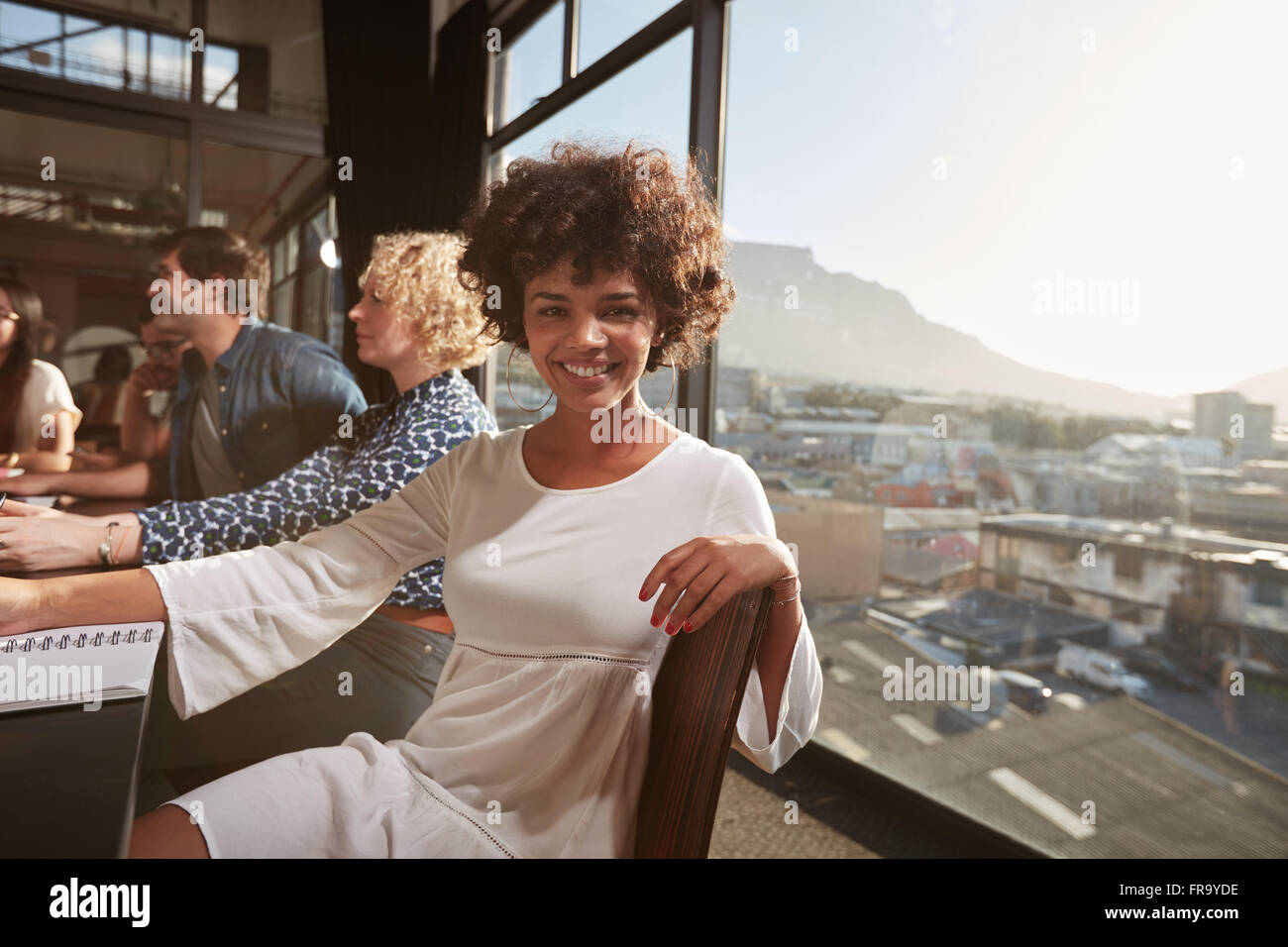 Porträt der glückliche junge Afrikanerin sitzen bei einem Treffen mit Kollegen im Hintergrund. Lächelnd, kreativ, professionell aussehende Stockfoto