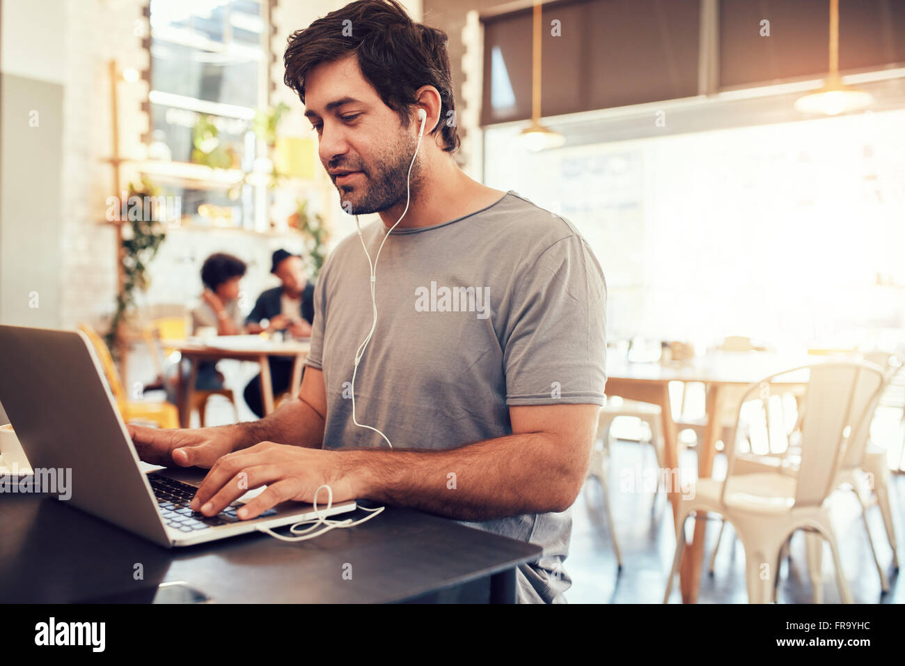 Porträt von hübscher junger Mann mit Kopfhörer am Laptop sitzen in einem Café arbeiten. Junger Mann an einem Café Surfen inte Stockfoto