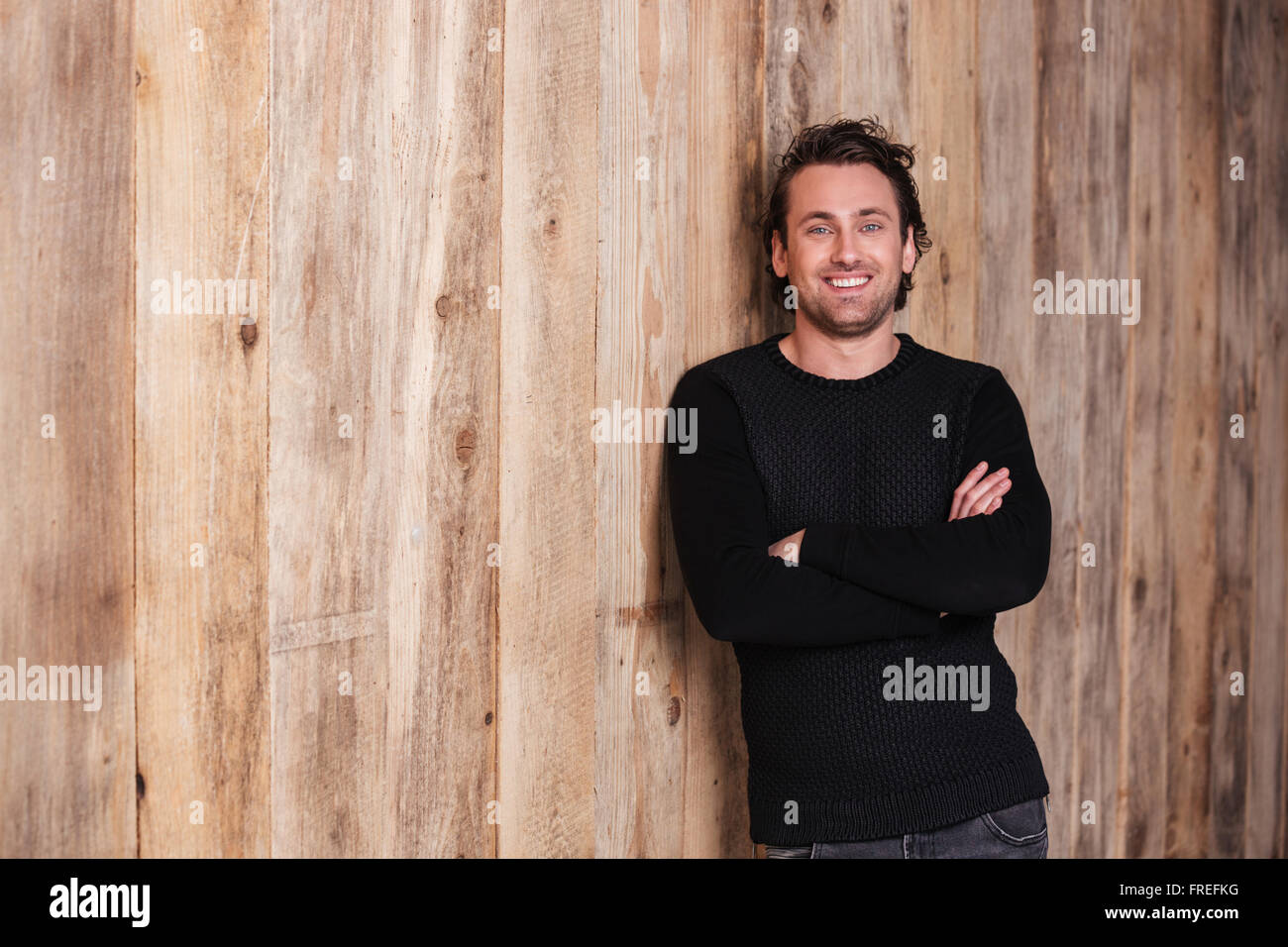 Lächelnder attraktiven junger Mann in schwarzer Pullover mit Händen stehend über hölzerne Hintergrund gefaltet Stockfoto