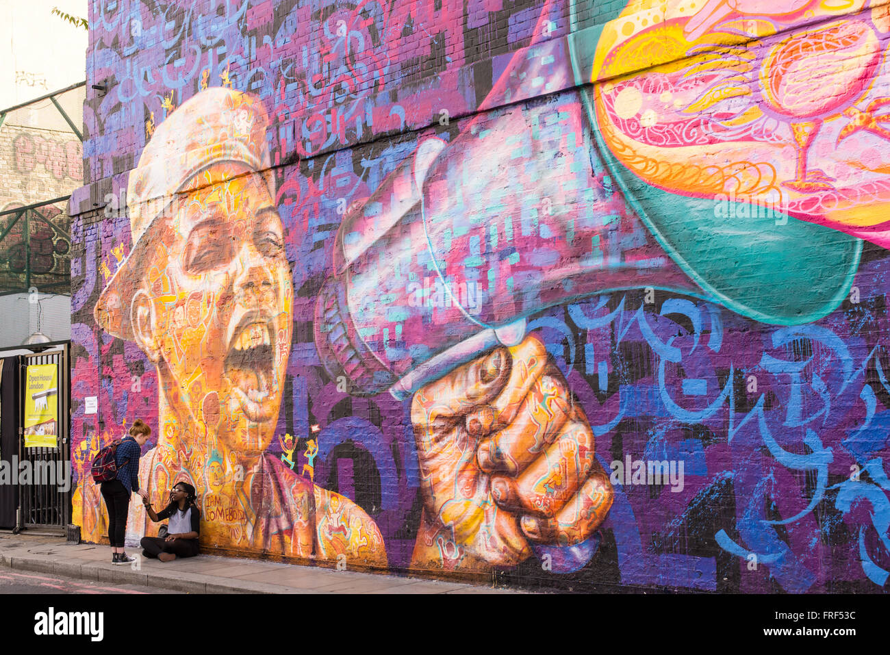 Drei Mädchen im Teenageralter stehen vor einer Wand mit einer großen Graffiti repräsentieren einen Jungen schreien mit einem Lautsprecher. Stockfoto