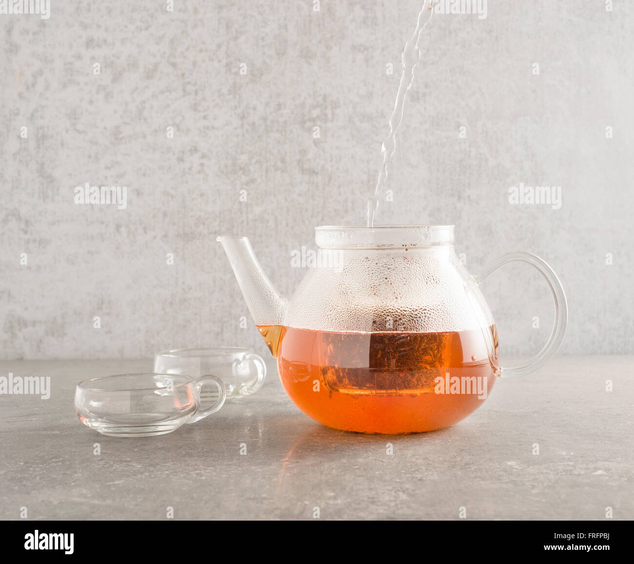 Heißes Wasser im Glas Teekanne gießen und Sichten Tee Blätter. Zwei Tassen aus Glas befinden sich auf der Seite auf einem Stein Küchentisch Stockfoto