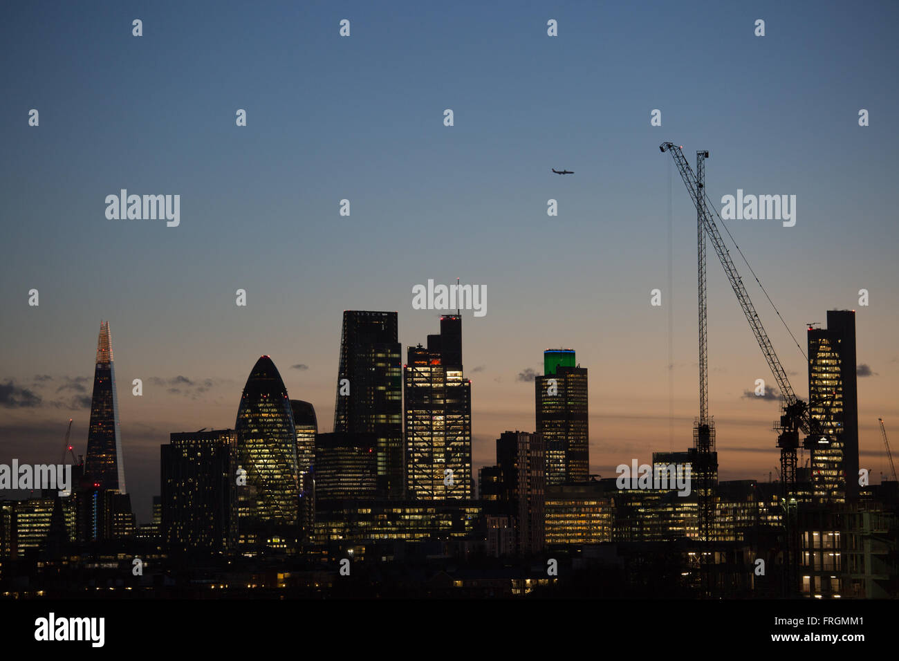 Die Sonne geht unter, auf der City of London und die Lichter sind an, gesehen von Hackney, East London. Eine Ebene verläuft in den Himmel. Stockfoto