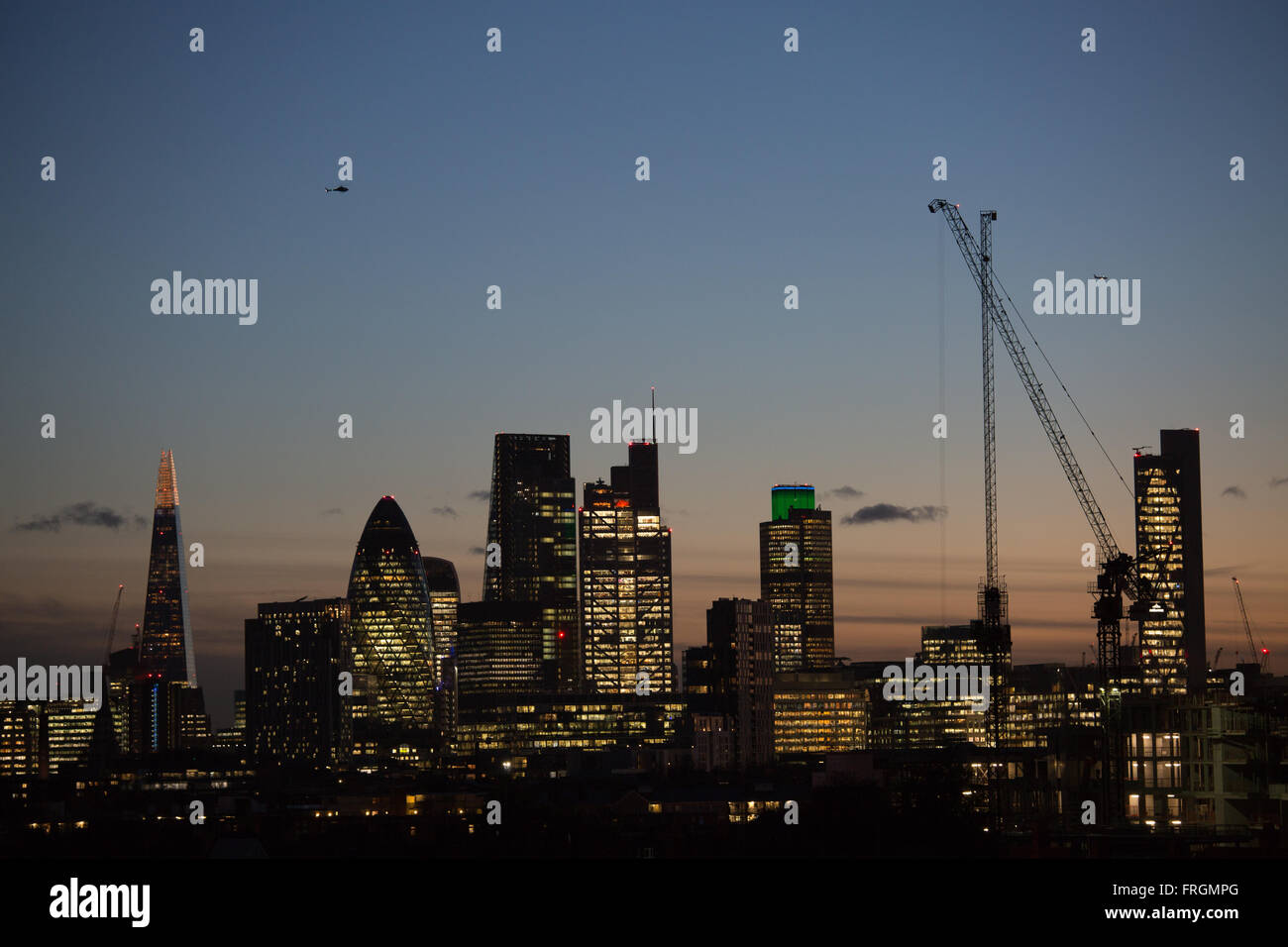 Die Sonne geht unter, auf der City of London und die Lichter sind an, gesehen von Hackney, East London. Ein Hubschrauber schwebt über. Stockfoto