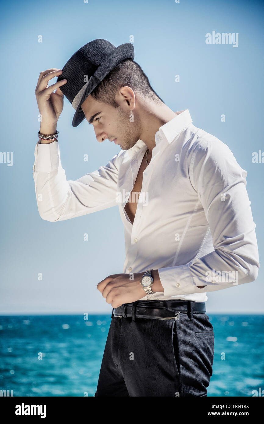 Schönen jungen Mann in elegantes weißes Hemd und schwarzen Filzhut, am Strand beim wegschauen. Wellen des Meeres im Hintergrund Stockfoto