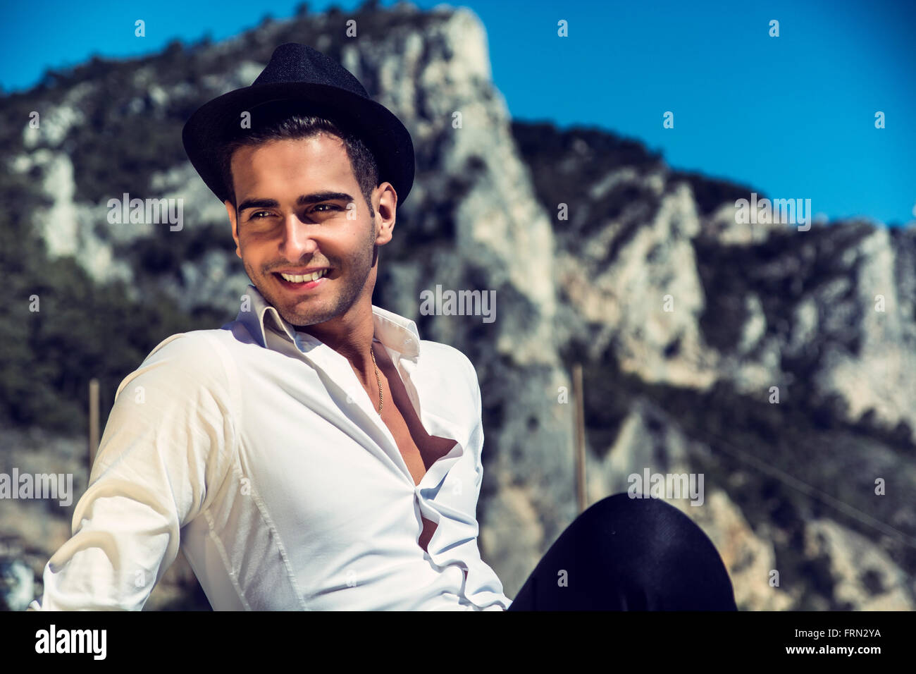 Schönen jungen Mann in elegantes weißes Hemd und schwarzen Filzhut, am Strand während aussehende lächelnd Weg. Hügel im Hintergrund Stockfoto