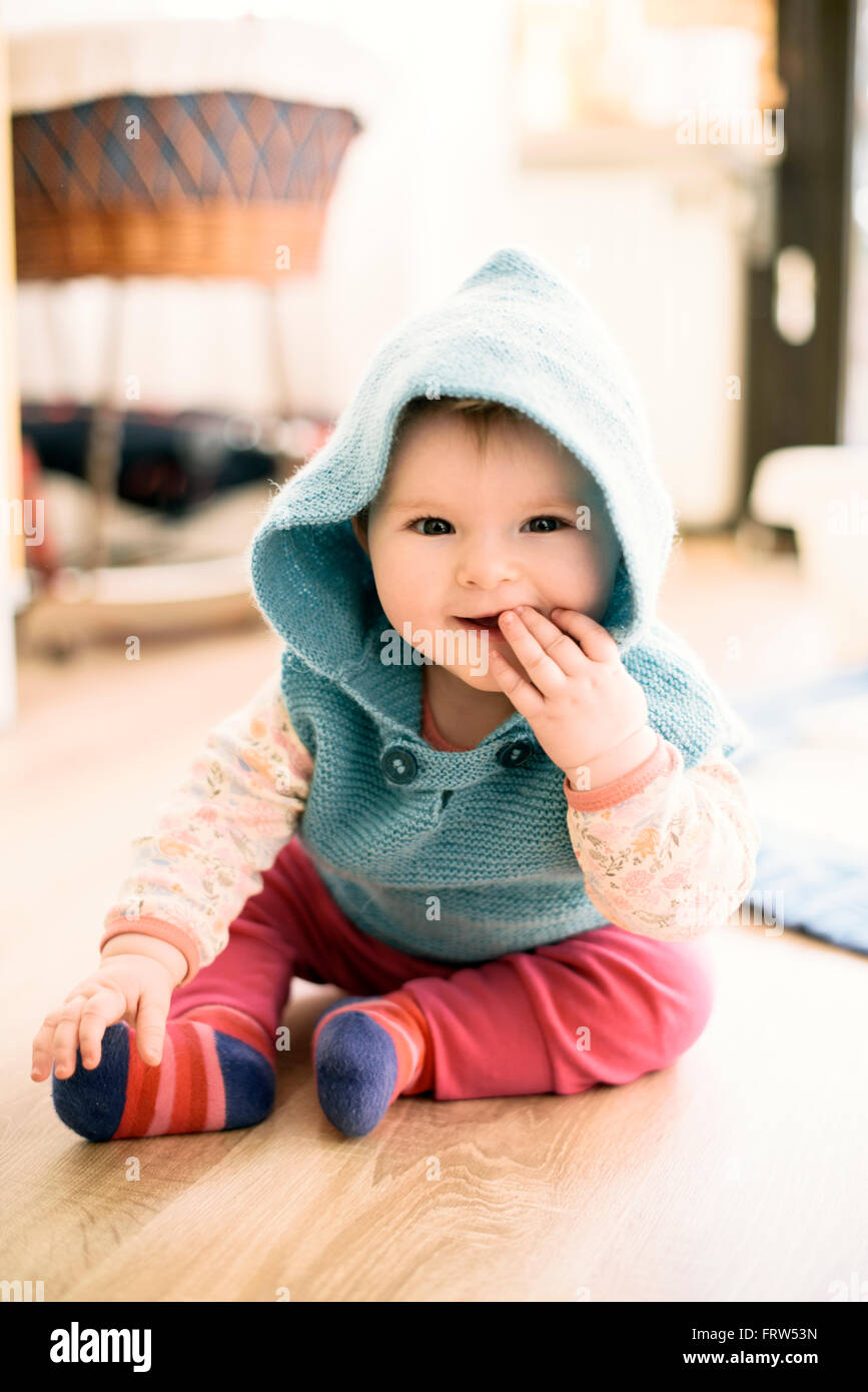 Porträt von lächelndes Babymädchen tragen Kapuzen Jacke sitzen auf dem Boden mit Finger im Mund Stockfoto