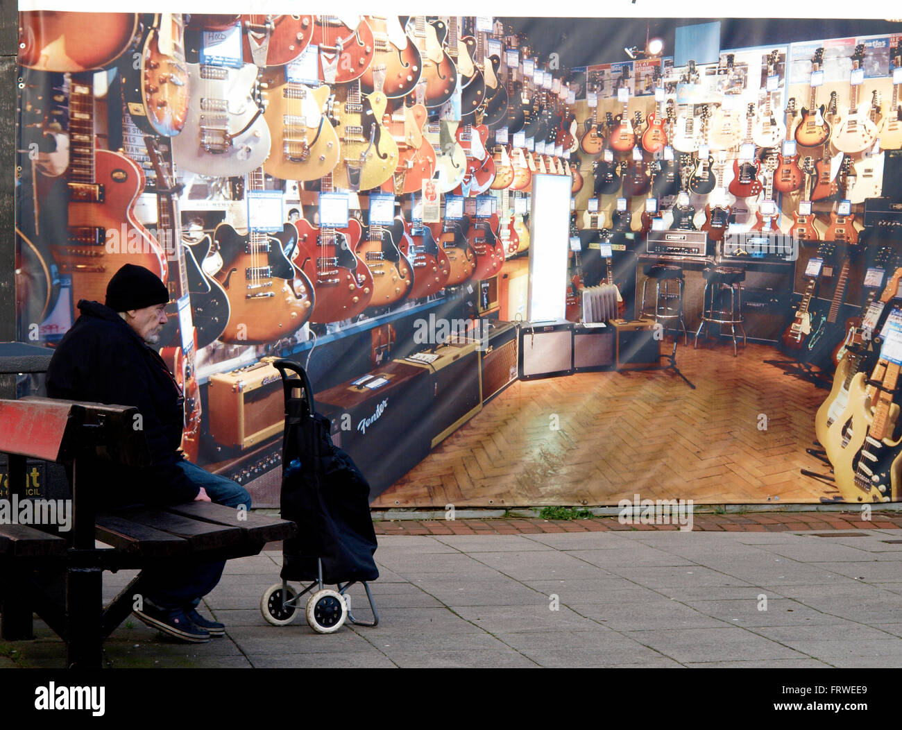 Alter Mann saß auf einer öffentlichen Bank vor einem bestiegen, Geschäft, bedeckt mit einem Foto des Innenraums von einem Musikgeschäft, Havant, Hants Stockfoto
