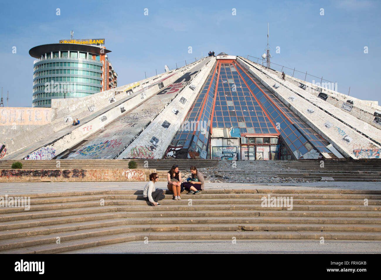 Leute sitzen auf den Stufen des Graffiti-bedeckten die Pyramide, das internationale Zentrum der Kultur mit Glas High-Rise Gebäude der österreichischen Raiffeisen Bank hinter, Tirana, Albanien Stockfoto