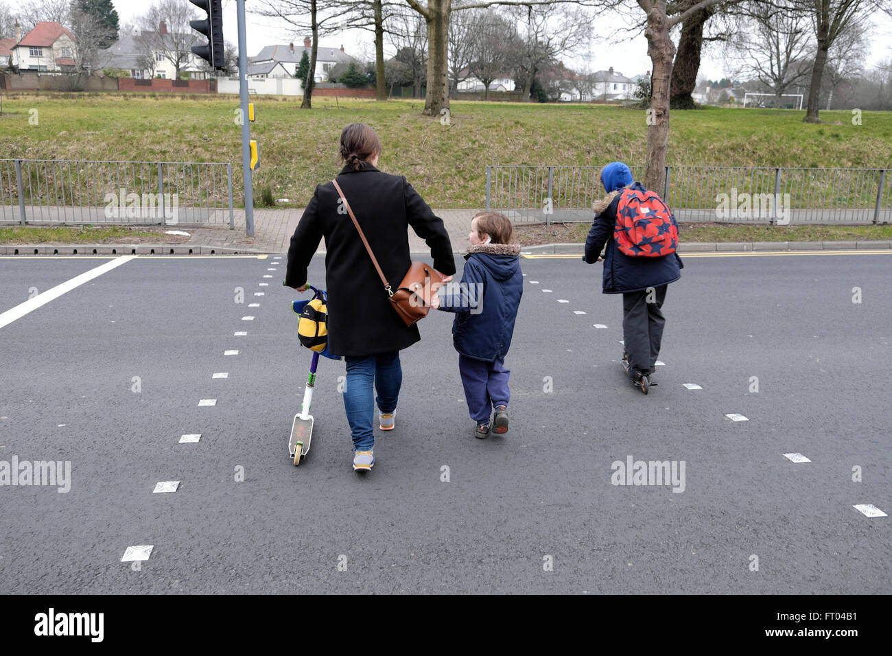 Mutter Wandern mit Kindern und Roller zur Schule Überqueren einer Straße an einem fußgängerüberweg, bevor es in Wales UK KATHY DEWITT zu arbeiten Stockfoto
