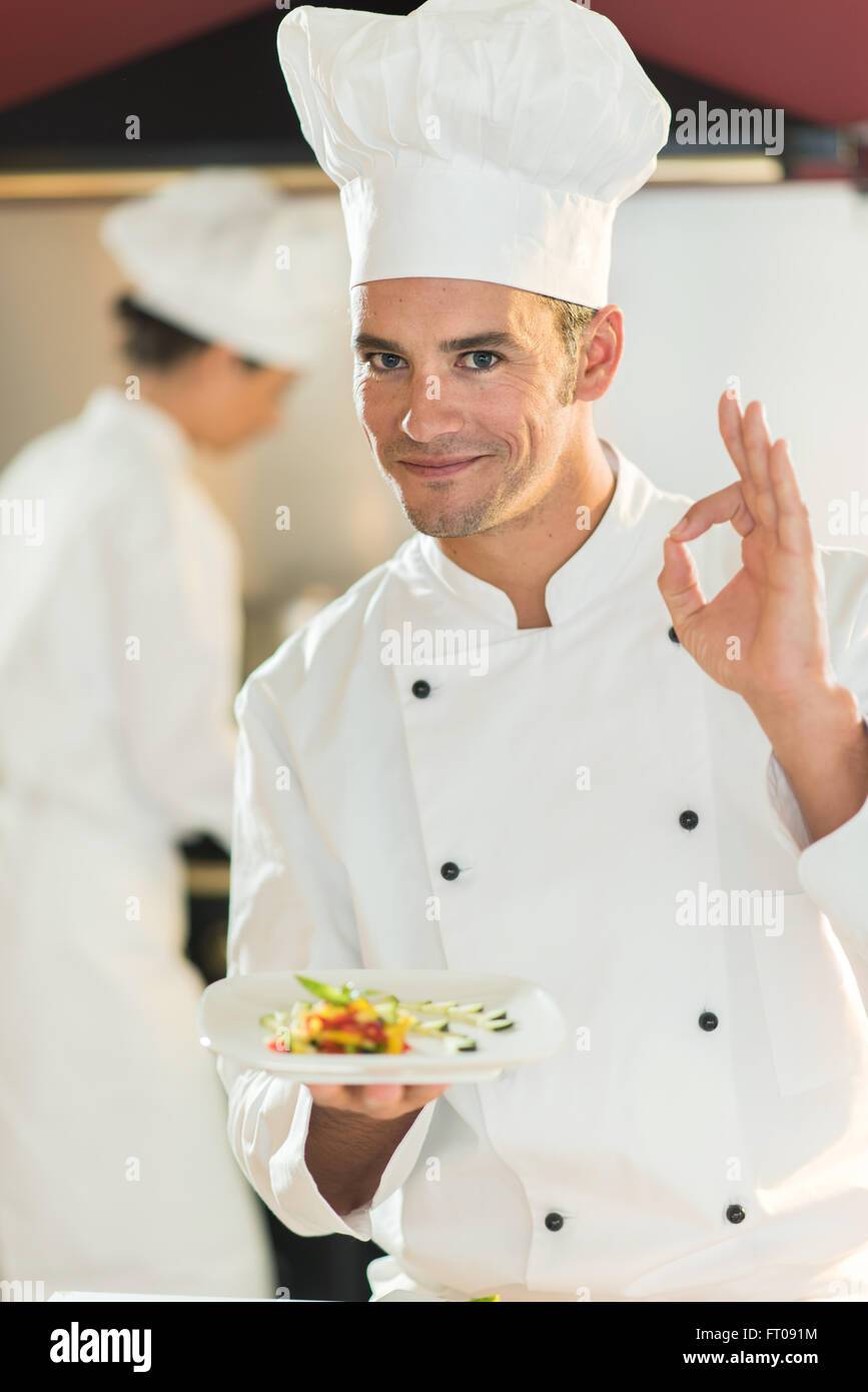 Ein Koch-Chef steht in einer professionellen Küche präsentiert einen Teller  mit feinem Essen. Er sucht nach Kamera tut die ok Zeichen tragen weiße Koch  Kleidung und Hut. Der Hintergrund jedoch unscharf Stockfotografie -
